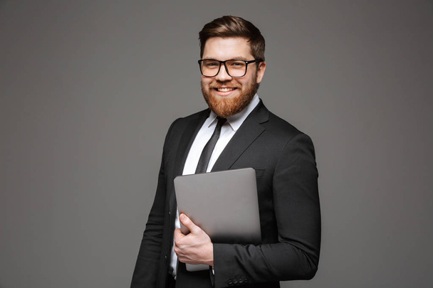 Portrait of a happy young businessman dressed in suit holding laptop computer and looking at camera isolated over gray background - Foto, imagen