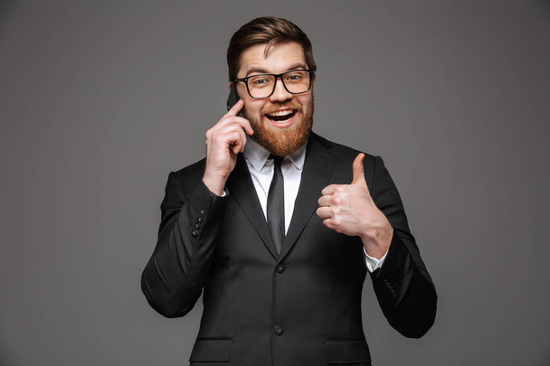Portrait of a happy young businessman dressed in suit talking on mobile phone and showing thumbs up isolated over gray background - Photo, Image