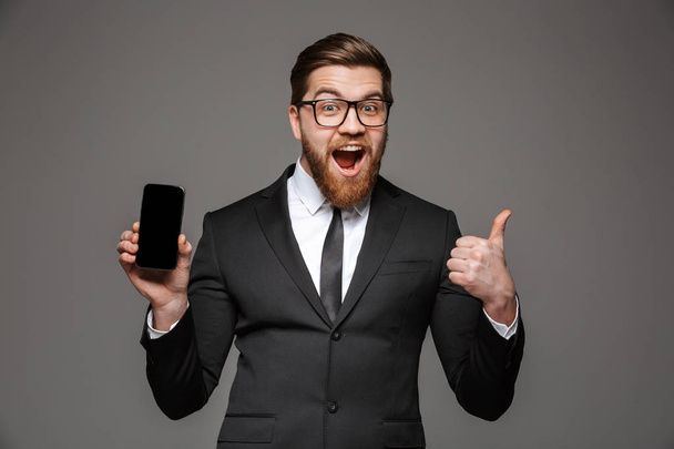 Portrait of a happy young businessman dressed in suit showing blank screen mobile phone and giving thumbs up isolated over gray background - 写真・画像