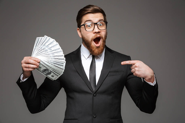 Portrait of a cheerful young businessman dressed in suit showing money banknotes isolated over gray background - Foto, Imagem