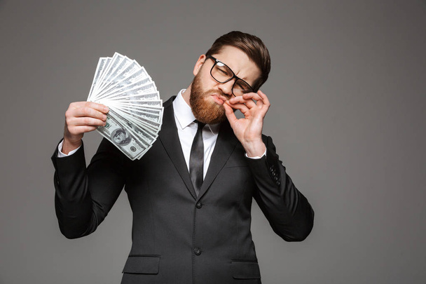 Porträt eines selbstbewussten jungen Geschäftsmannes im Anzug, der Geldscheine isoliert vor grauem Hintergrund zeigt - Foto, Bild