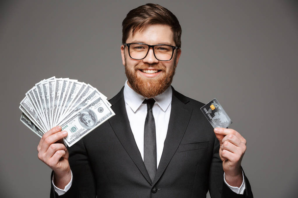 Retrato de un joven empresario feliz sosteniendo billetes de dinero y tarjeta de crédito aislados sobre fondo gris
 - Foto, imagen