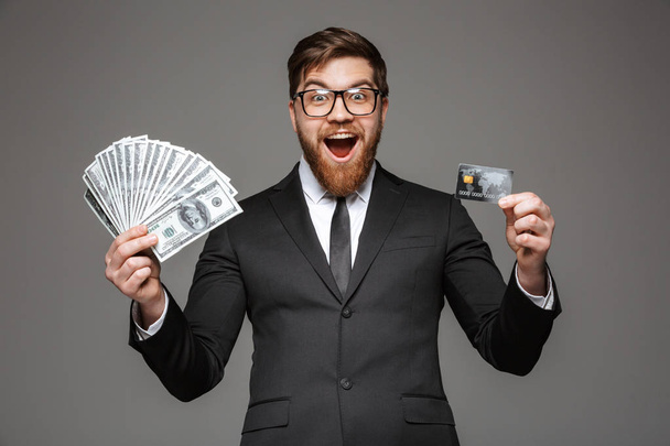 Portrait of an excited young businessman holding money banknotes and credit card isolated over gray background - Valokuva, kuva