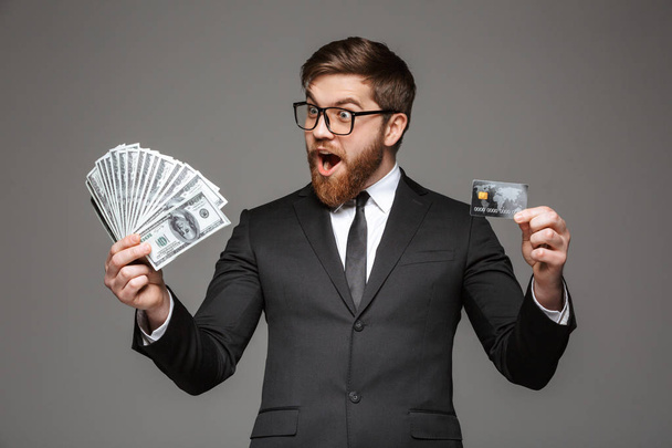 Portrait of a surprised young businessman holding money banknotes and credit card isolated over gray background - Fotó, kép