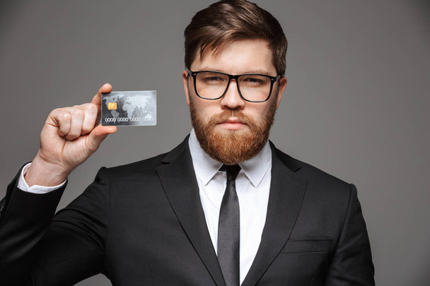 Portrait of a happy young businessman showing credit card isolated over gray background - Fotografie, Obrázek