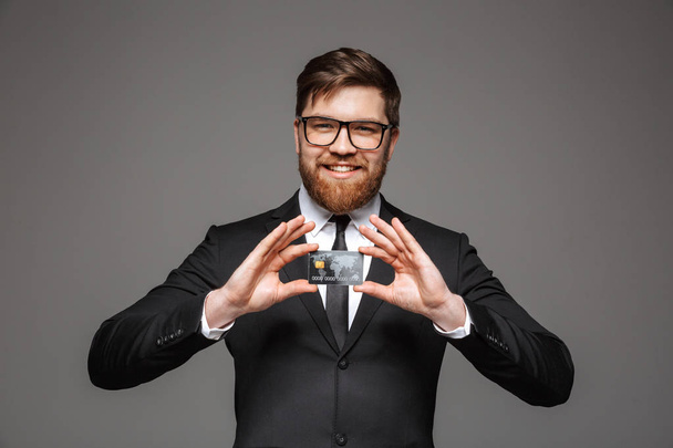 Portrait of a happy young businessman showing credit card isolated over gray background - Fotografie, Obrázek