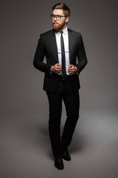 Full length portrait of a confident young businessman dressed in suit posing while standing and looking away isolated over gray background - Foto, afbeelding