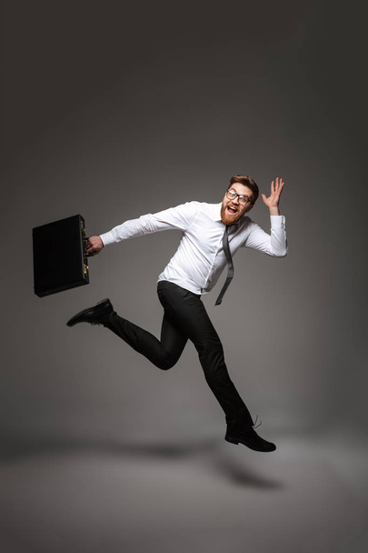 Full length portrait of an excited young businessman posing while holding briefcase and jumping isolated over gray background - Foto, imagen