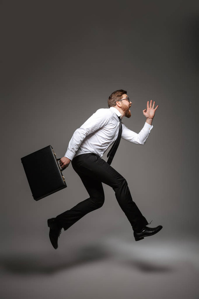 Full length portrait of a happy young businessman posing while holding briefcase and jumping isolated over gray background - Foto, Bild