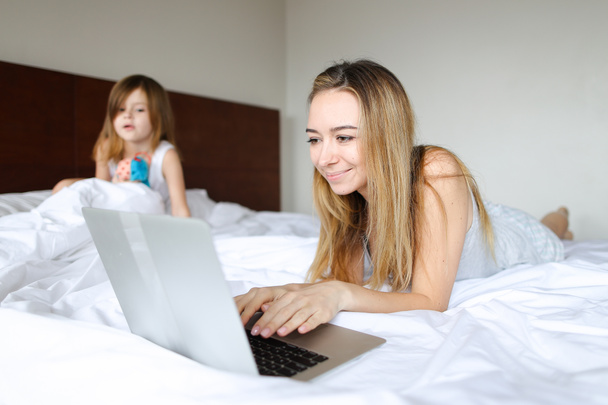 Молодая мать лежит в кровати с маленькой дочкой и пользуется ноутбуком
. - Фото, изображение