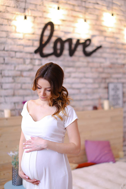 Mulher europeia grávida vestindo vestido branco segurando barriga, amor inscrição na parede de tijolo
. - Foto, Imagem