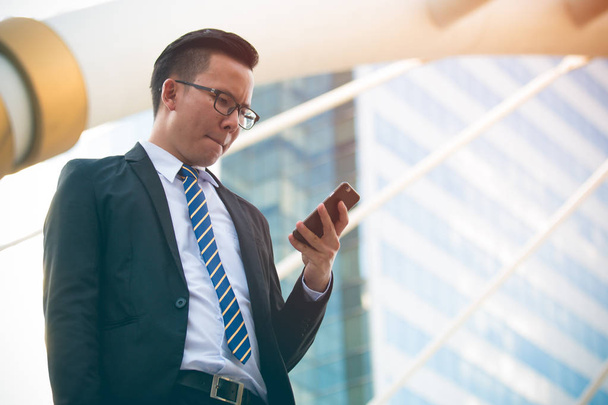 Moderni nuori liikemies käyttää musta puku käsi tilalla älypuhelin. Ammattimainen liikemies seisoo toimiston ulkopuolella ja etsii matkapuhelinta, jolla on vakava ilme. kopiotilaa
 - Valokuva, kuva