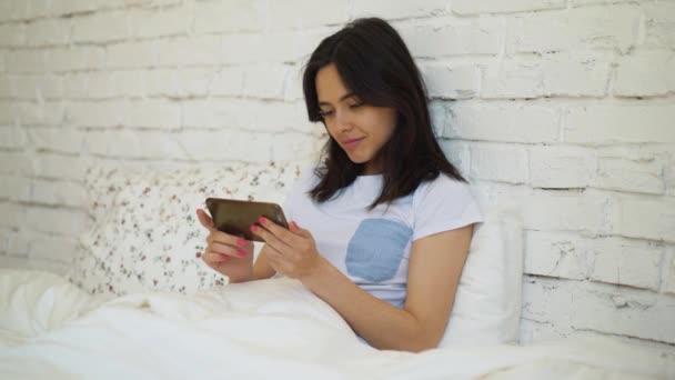 Side view of young woman lying in bed and video chatting using her smartphone - Felvétel, videó