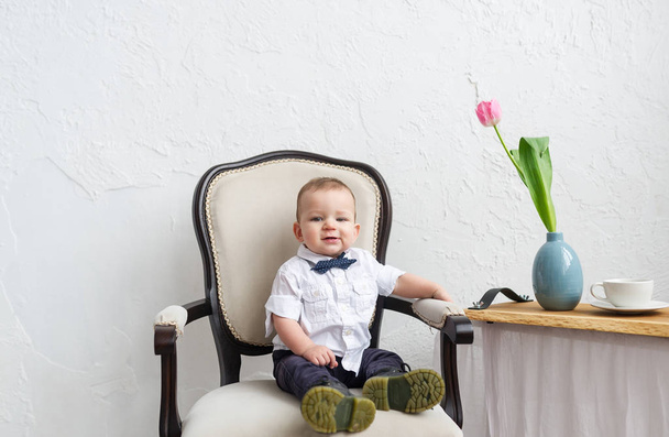 Niño de moda sentado en el sillón en la habitación blanca. Retrato horizontal
 - Foto, Imagen