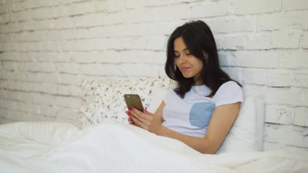 Vista lateral de la mujer joven acostada en la cama y video chat con su teléfono inteligente
 - Metraje, vídeo