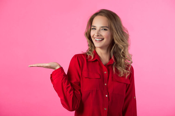 young joyful beautiful girl in red attire on a pink background with a gesture of hands - Фото, изображение
