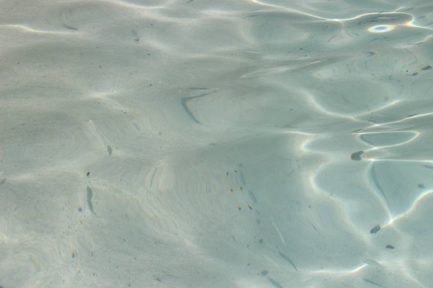 Αμμώδης παραλία με ρηχά νερά με διαφανή νερό, στην επιφάνεια των οποίων φως του ήλιου και κύματα δημιουργούν ένα μοτίβο. Μια σαφή άποψη για την αμμώδη βυθό με σκιές από επιπλέοντα ψάρια. Απαλή εστίαση. - Φωτογραφία, εικόνα