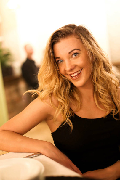 Портрет красивой счастливой улыбающейся девушки в черном платье, сидящей в ресторане на свидании в ожидании блюда, смеющейся
 - Фото, изображение