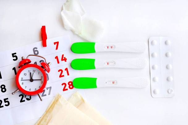 Tira de prueba de ovulación con reloj rojo calendario, píldoras anticonceptivas y almohadilla menstrual. Las pruebas muestran tres resultados diferentes-Resultado positivo, Es un momento para el bebé. Días estériles. La prueba no está bien hecha.
 - Foto, imagen