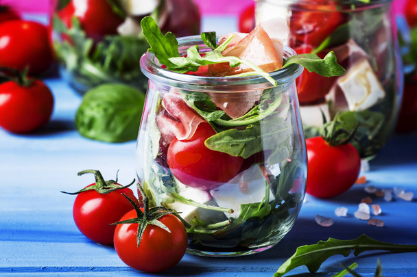 Salad with arugula, cheese, cherry tomatoes and prosciutto in glass jars, pink blue background, selective focus - Photo, image