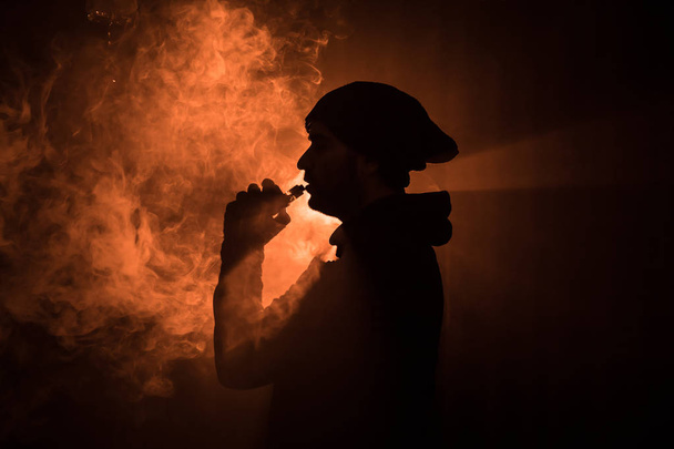 Vaping man holding a mod. A cloud of vapor. dark foggy toned background of clouds of smoke. Vaping an electronic cigarette with a lot of smoke. Vape concept. Selective focus - 写真・画像