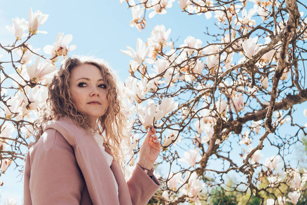curly elegant woman in pink coat posing next to blooming fragrant magnolia in the park - Foto, afbeelding