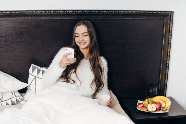 laughing long-haired girl in white pajamas sits in bed early in the morning, looks into her smartphone and does not want to get up - Valokuva, kuva