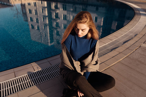 lovely blonde girl in a stylish blue sweater sits by the pool outdoors and resting - Foto, afbeelding