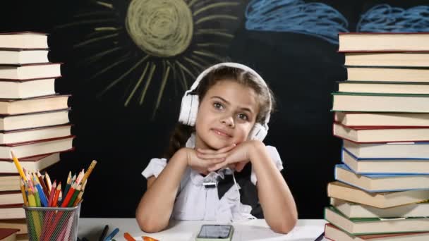 Moderne school-concept. Aantrekkelijk meisje zit achter een bureau met stapels boeken. en een schoolbord achter haar. Schoolmeisje in hoofdtelefoons glimlacht. - Video
