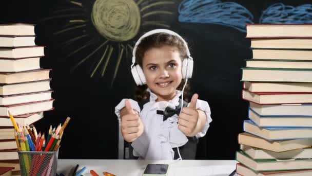 Modern school concept. Attractive girl sits at a desk with heaps of books. and a blackboard behind her. positive schoolgirl in headphones smiles. thumbs up. - Footage, Video
