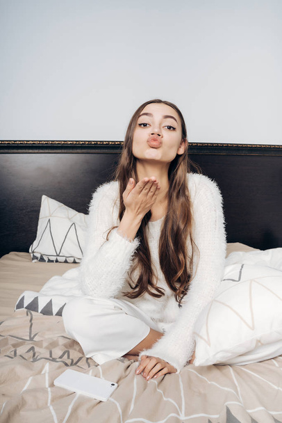 cute long-haired girl in white pajamas sits on the bed early in the morning, sends an air kiss - Foto, Bild