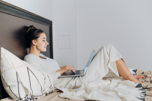 cute young girl in white pajamas sitting in bed and watching an interesting movie on her laptop - Fotó, kép