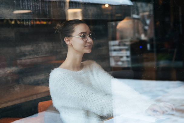 beautiful young freelancer girl in white sweater and glasses sitting in a cafe thinking about work, smiling - Fotoğraf, Görsel