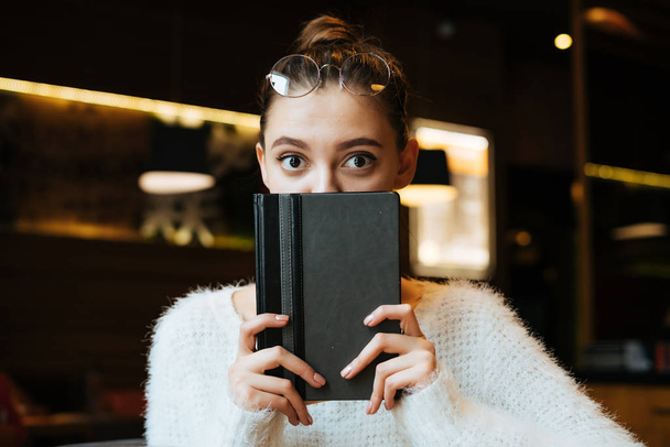cute funny girl student in a white jacket covers half of his face with a notebook, sitting in a cafe and looking at the camera - Foto, Imagen