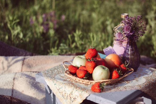 maçãs e morangos em uma chapa de vime, flores roxas em um vaso de vidro, em um banquinho de madeira com um guardanapo decorativo
 - Foto, Imagem