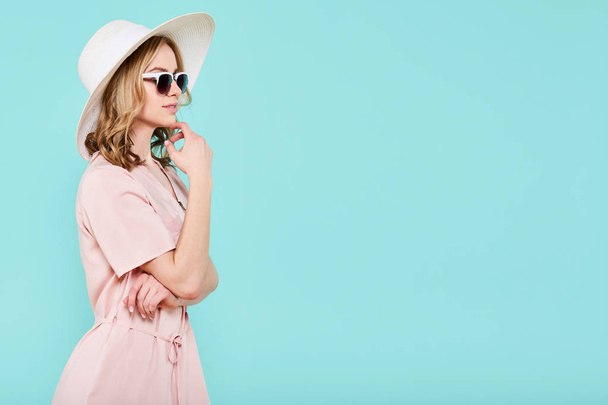 Elegant young attractive woman wearing pale pink summer dress, straw hat and sunglasses, thinking about her summer vacation. Side view of woman with hand on chin, isolated over pastel blue background. - Фото, изображение