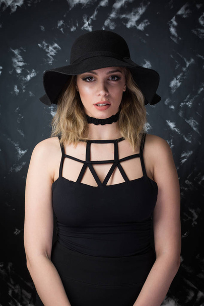 Modern young woman in black dress and wide brim hat posing. No retouch, studio lighting, closeup - Foto, imagen