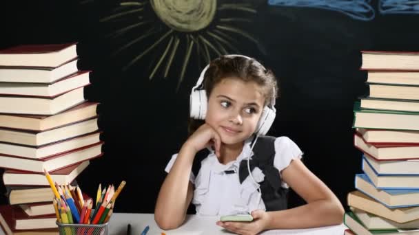 Concept van de school. Aantrekkelijk meisje zit achter een bureau met stapels boeken. en een schoolbord achter haar meisje luistert naar muziek - Video