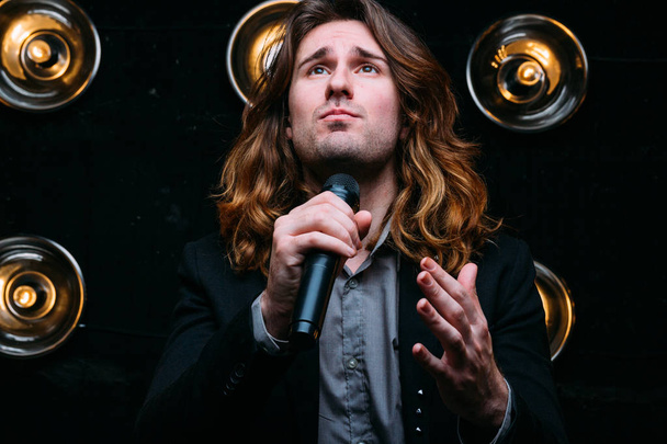 Brutal singer with microphone on the stage, with long hair, against the background of bright lanterns, scene - Photo, image