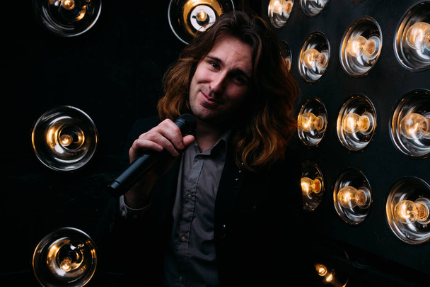 Brutal singer with microphone on the stage, with long hair, against the background of bright lanterns, scene - Photo, Image