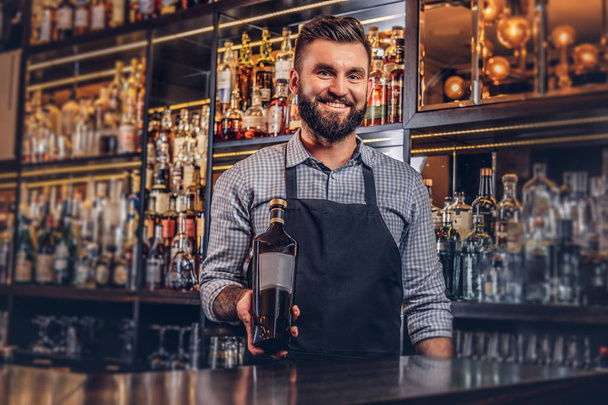 Vrolijke stijlvolle brute barman in een shirt en schort presenteert een fles exclusieve alcohol bar teller achtergrond. - Foto, afbeelding