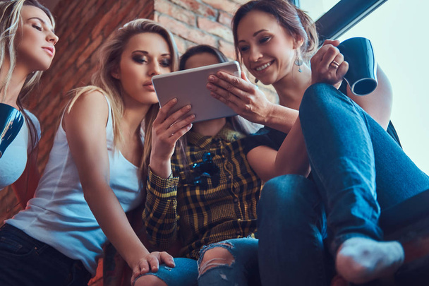 Grupo de amigas em roupas casuais discutindo enquanto procuravam algo em um tablet digital em um quarto com um interior loft
. - Foto, Imagem