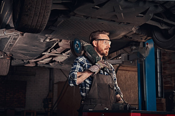 Automechaniker in Uniform und Schutzbrille hält einen Winkelschleifer in der Hand, während er in einer Werkstatt unter dem Hubwagen steht.  - Foto, Bild