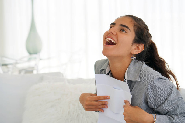 Young woman enjoying good news in writing. The girl reads a letter with good news sitting on the couch. An euphoric girl is happy after reading good news in a written letter, approving a loan. - Photo, Image