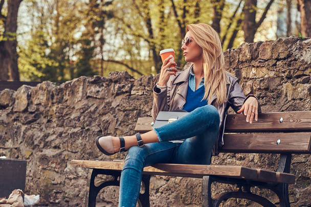 Fashionable blonde female relaxes outdoor, drinking takeaway coffee while sitting on the bench in city park. - Photo, Image