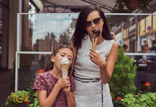 Fashionable mother and cute daughter enjoy ice cream on a hot summer day, standing near a cafe. - Photo, Image