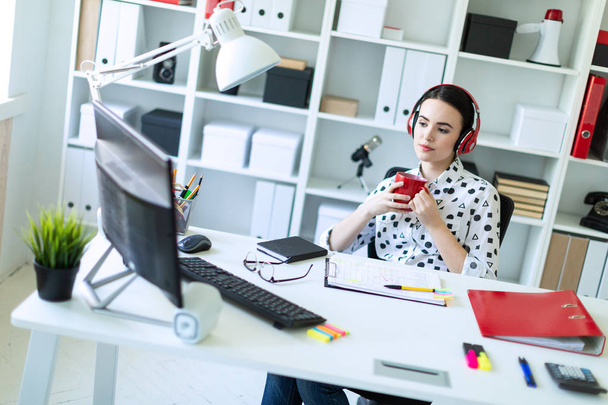 Młoda dziewczyna siedzi w słuchawki przy stole w biurze, w jej ręce trzyma kubek czerwony i wygląda na monitor. - Zdjęcie, obraz