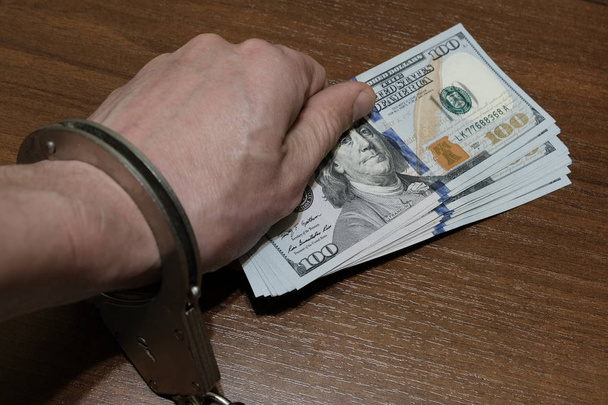 Ręka mężczyzny w kajdankach ustanawia paczkę dolarowe na powierzchnię stołu w kolorze orzech. Koncepcja naruszenia prawa, korupcji, nadużyć finansowych. - Zdjęcie, obraz