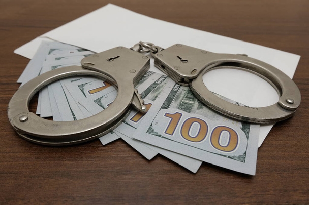 Concept violation of the law, corruption, financial fraud. The handcuffs lie on an envelope in which are many hundred dollar bills. Brown wooden background. - Photo, Image