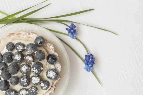 Fresh blueberry tartlet with vanilla custard in portion plate close-up, next to elegant blue flowers, , white napkin. Delicious and romantic summer berry dessert, copy space. Selective focus - Fotografie, Obrázek